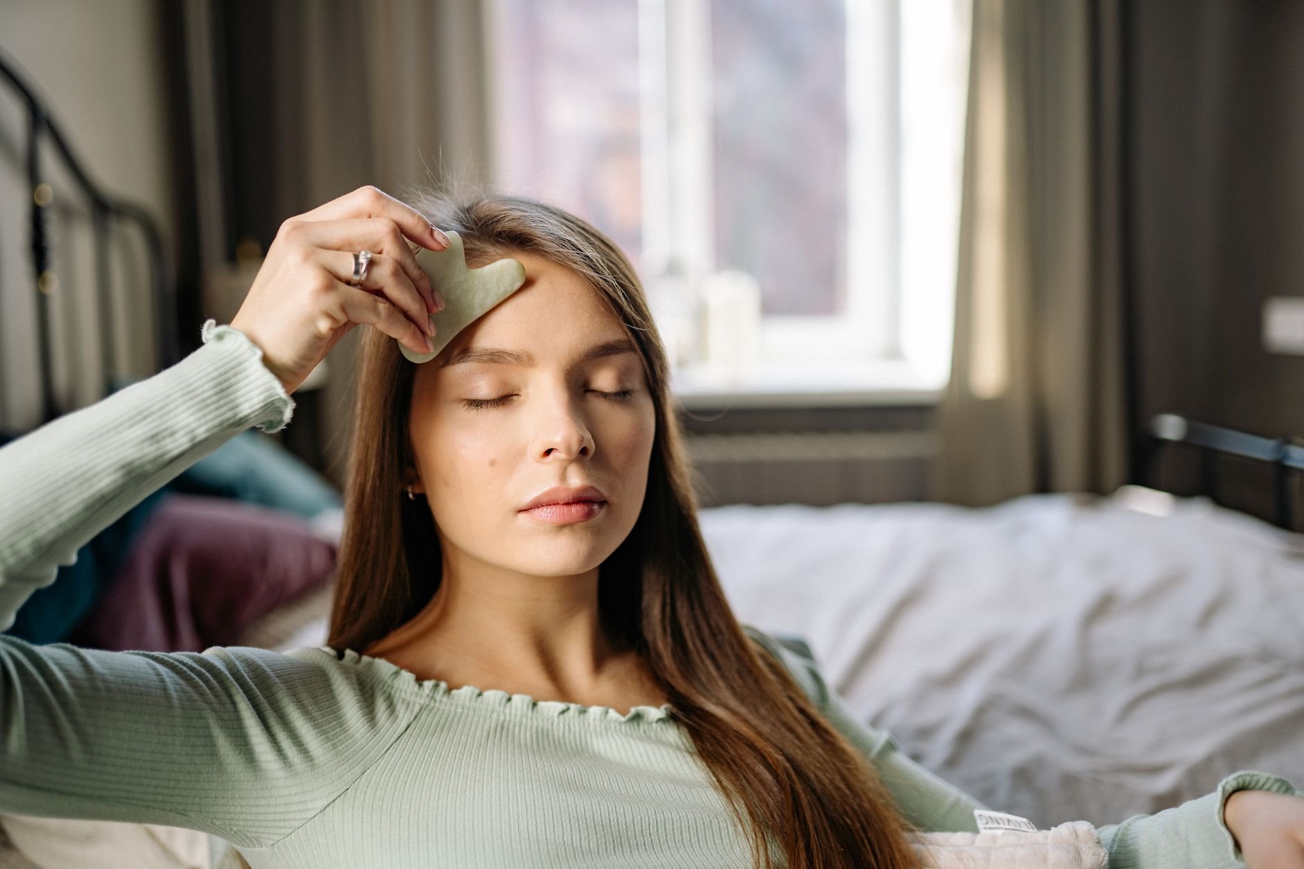 Woman using a gua sha stone during a morning skincare routine, eyes closed, relaxed indoors.